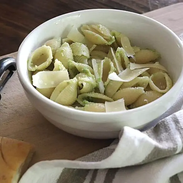 Σκόρδο scape pesto recipe image που δείχνει μπολ με κέλυφος σχήμα ζυμαρικών και σκόρδο scape pesto που στηρίζεται σε ένα ξύλινο σκάφος με μια ριγέ καφέ και λευκή πετσέτα που βρίσκεται δίπλα στο μπολ